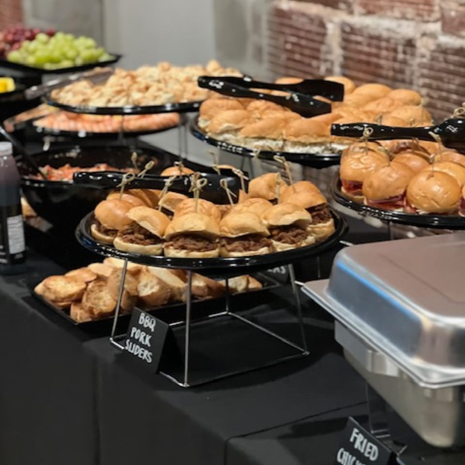 A catering spread with BBQ pork sliders, other sandwiches, and fruit on a black tablecloth.