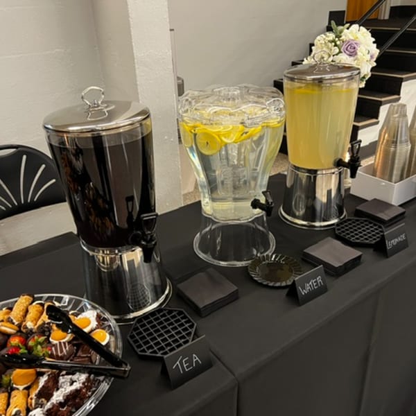 A catering table with dispensers for tea, lemon water, and lemonade, alongside a dessert platter.