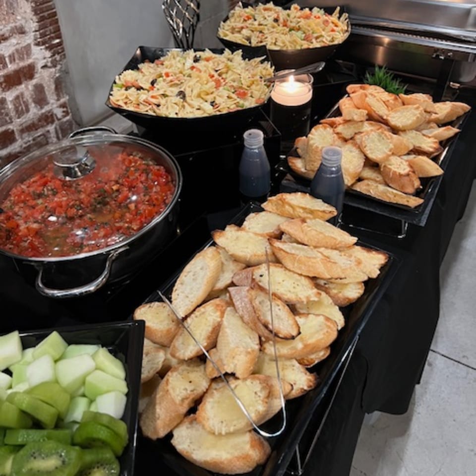 A buffet spread with pasta salad, bruschetta, a pot of tomato sauce, and fruit.