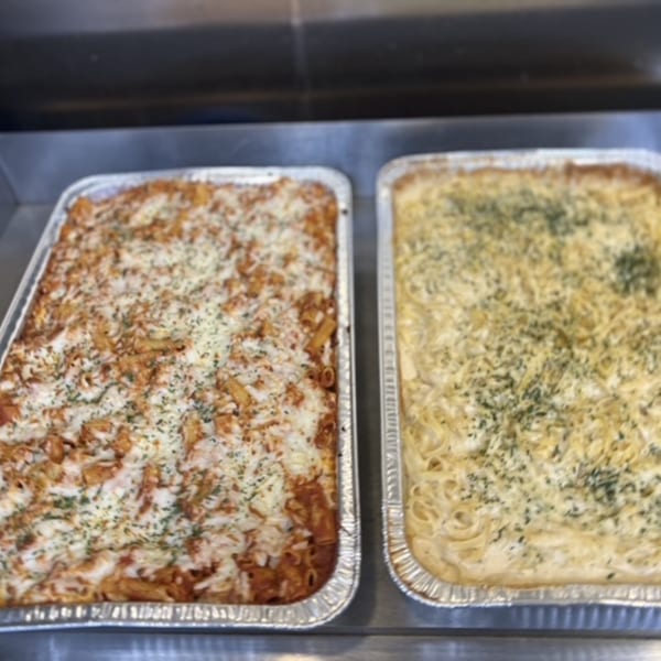 Two large catering trays of baked pasta, one red sauce and one white sauce, topped with cheese and herbs.