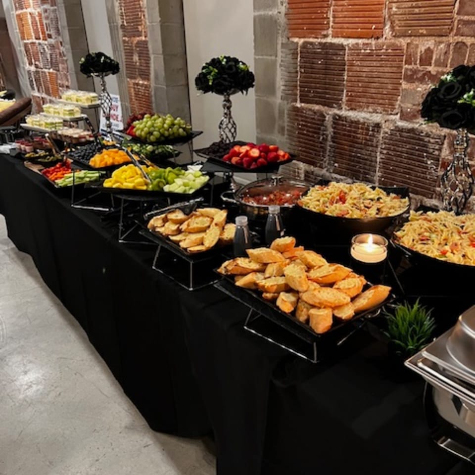 A buffet table with fruit, pasta, bread, and desserts, set against a brick wall.