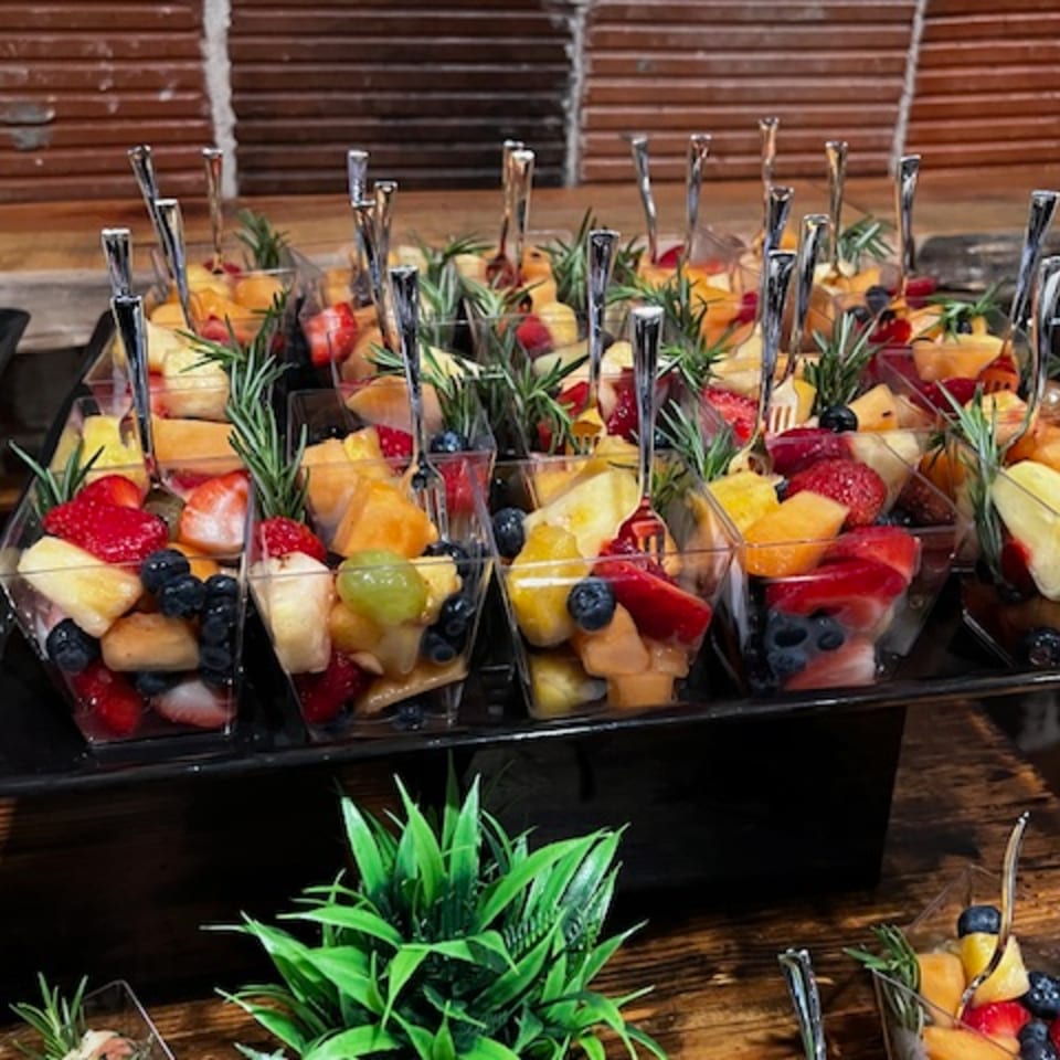 Rows of fruit cups with strawberries, blueberries, melon, and pineapple, garnished with rosemary.