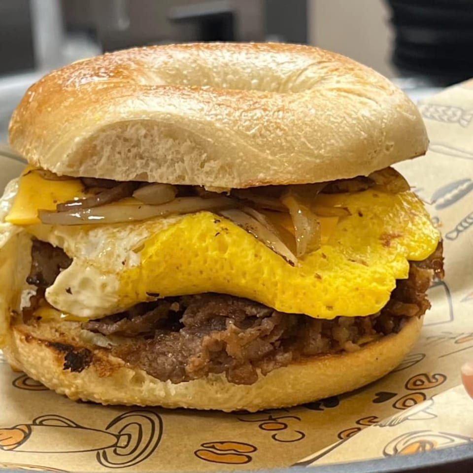A breakfast bagel with egg, cheese, sausage, and caramelized onions on branded paper.