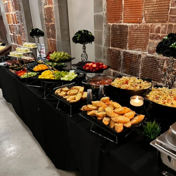 A buffet table with various fruits, pasta salads, and bread against a brick wall.