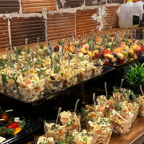 Rows of individual servings of pasta salad and fruit salad in clear cups on a buffet.