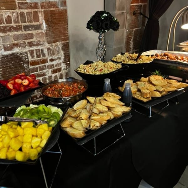 A buffet spread with various dishes including fruit, pasta, salsa, and bread against a brick wall.
