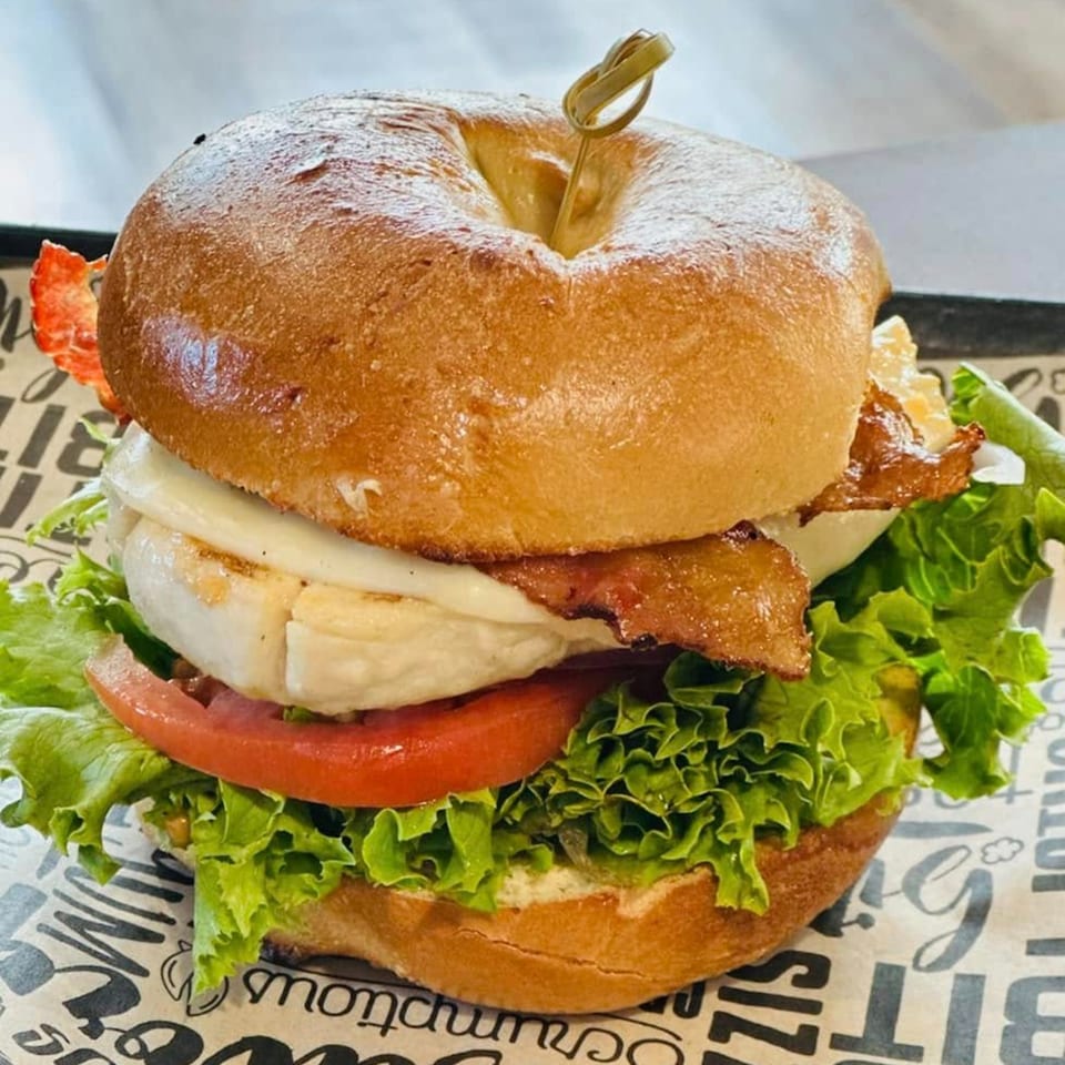 A bagel sandwich with grilled chicken, bacon, cheese, lettuce, and tomato on a branded paper.