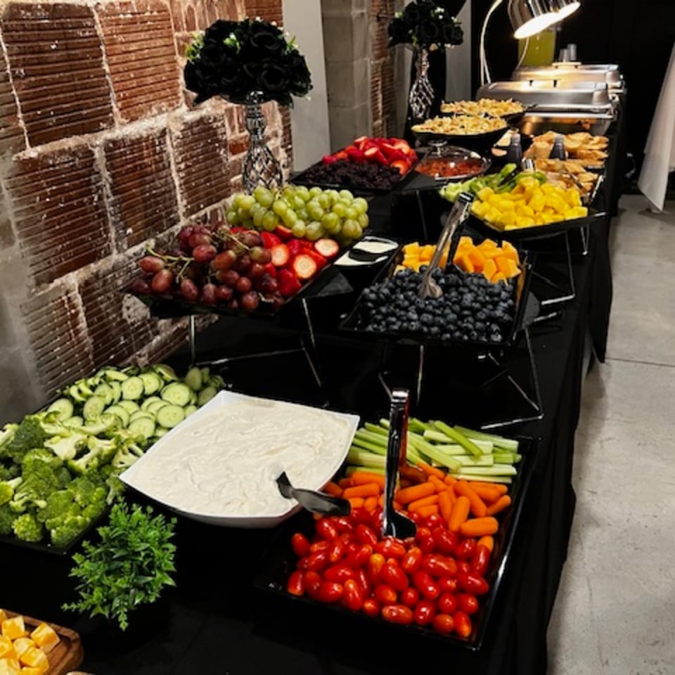 A long buffet table laden with fresh fruit, vegetables, and dips, with chafing dishes in the background.