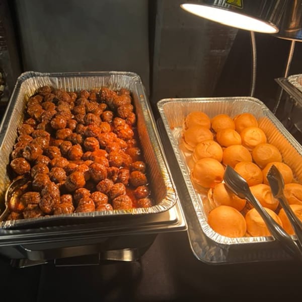 Catering trays of meatballs in sauce and dinner rolls with tongs under a heat lamp.