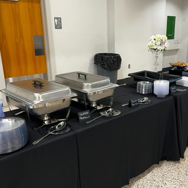 Catering buffet with two chafing dishes, serving utensils, and disposable plates on a black-draped table.