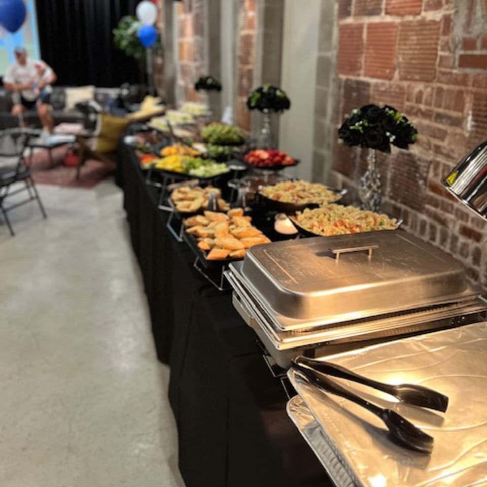 A long buffet table with various dishes, including pasta, bread, and salads, at Mary's Bagel Cafe.